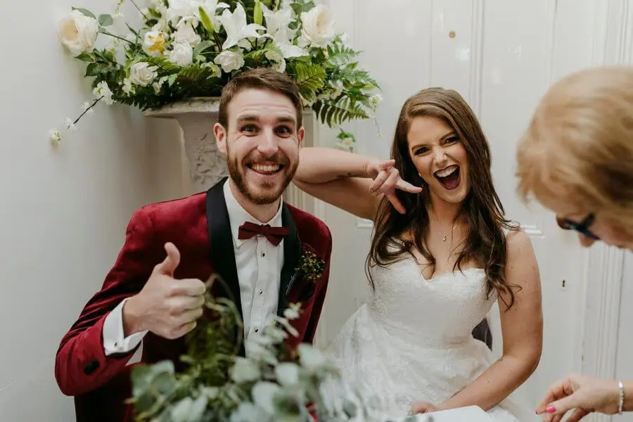 Bride and groom laughing beside their signing table as he raises a thumb.