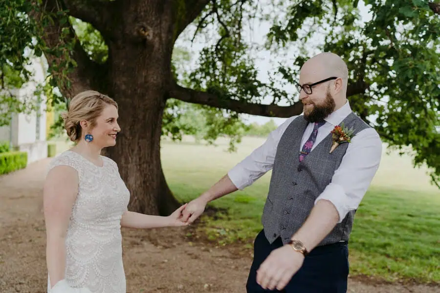 Bride and groom holding hands and smiling beneath a large tree.