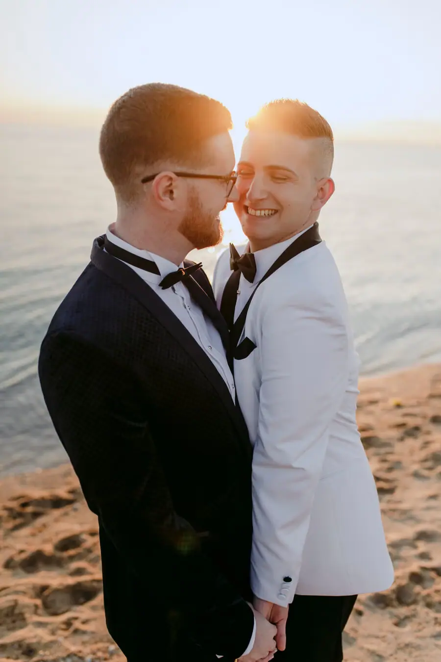 Two grooms embracing on a beach in golden sunset light.