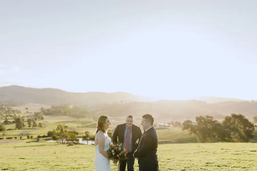 Couple and a suited man standing on a sunlit hillside.