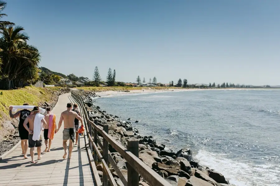 People walking along a seaside boardwalk beside rocks and calm water.