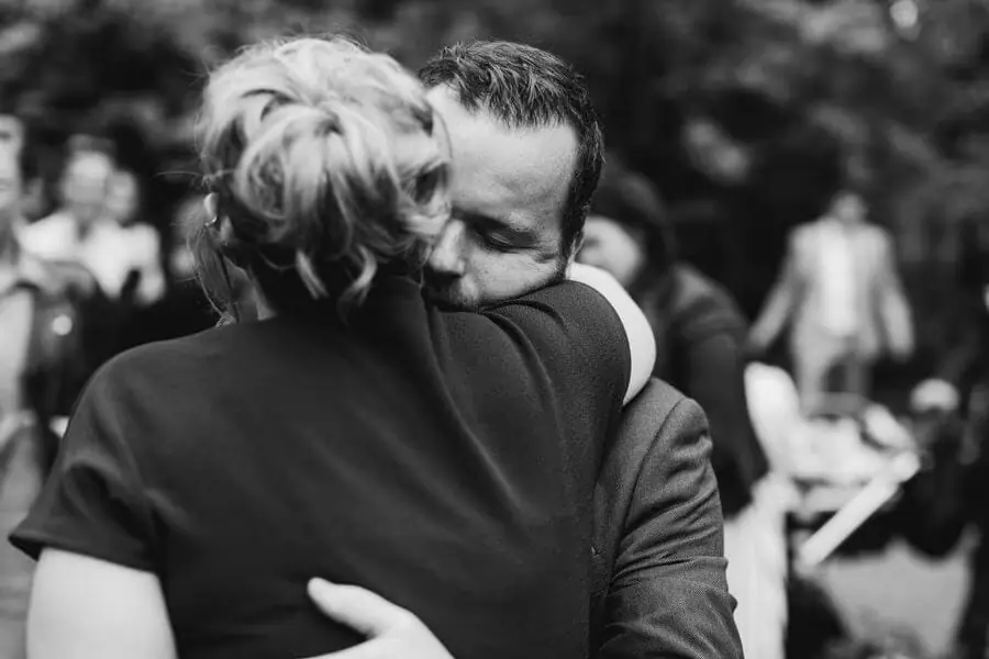 Black-and-white photograph of a man hugging a woman among guests outdoors.