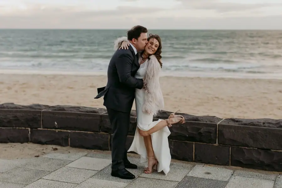 Couple embracing beside a seaside wall as the bride lifts one leg.