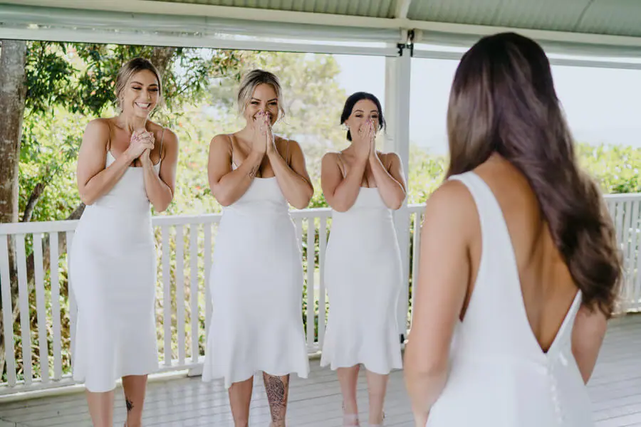 Three women in white dresses reacting to the bride on a veranda.