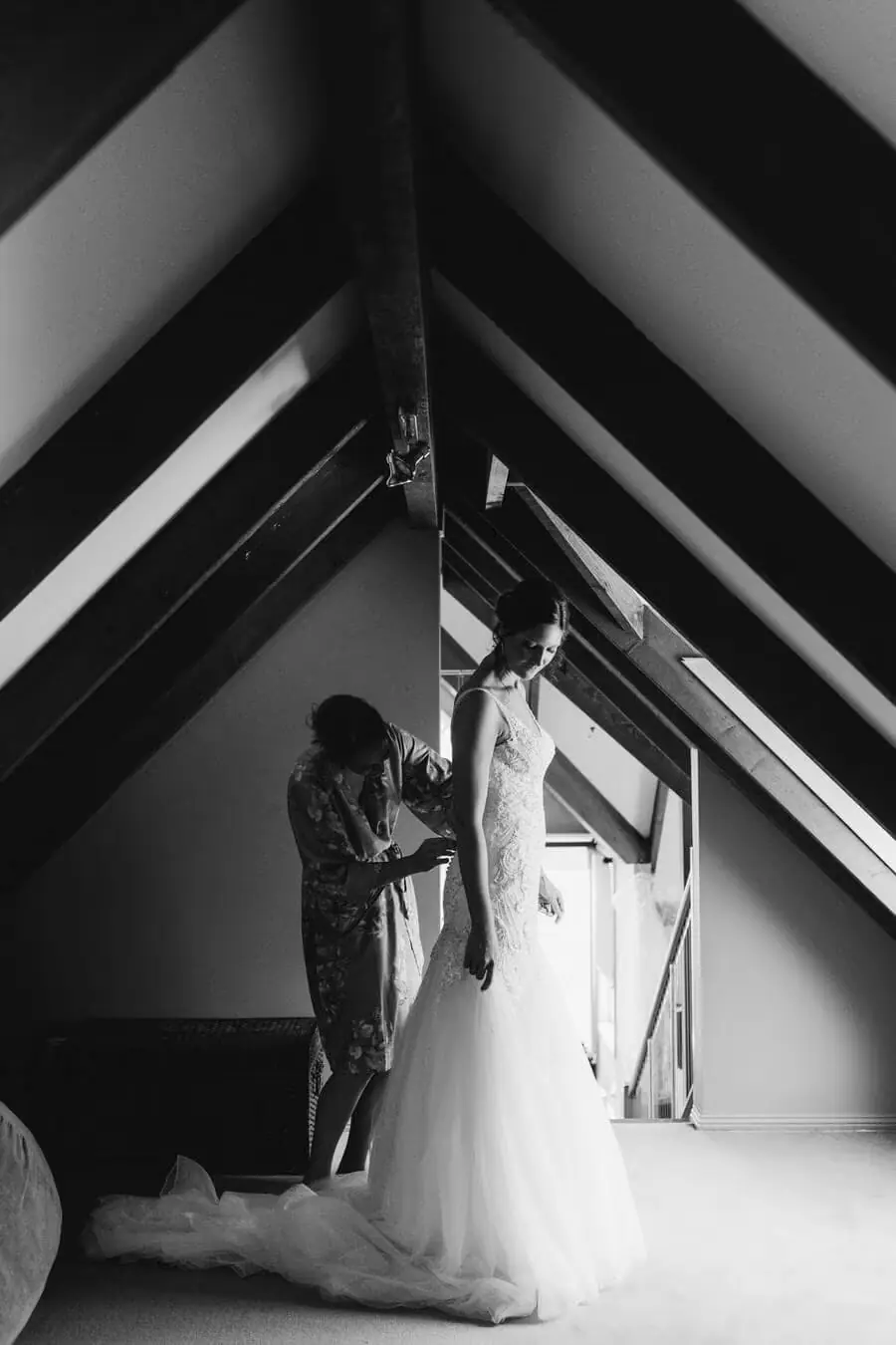 Black-and-white photograph of a woman helping the bride dress in a loft room.