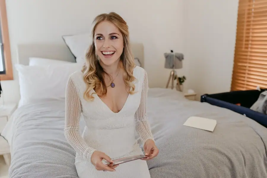 Bride smiling while holding a small card beside a bed.