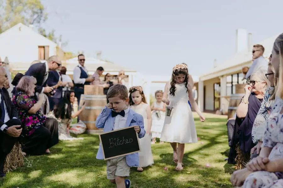 Children walking down an outdoor aisle as a boy carries a chalkboard sign.