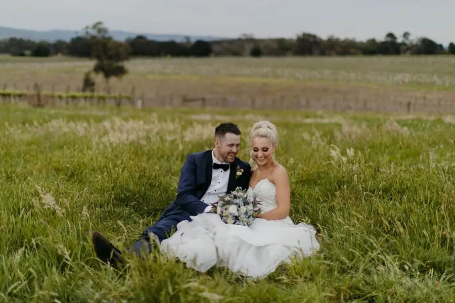 Bride and groom laughing together while sitting in tall green grass.