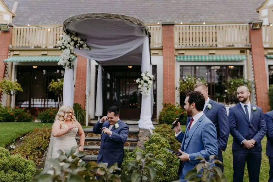 Couple and companions laughing outside a brick building with a draped ceremony arch.