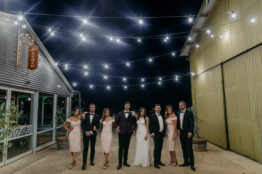 Wedding party walking between buildings beneath string lights at night.