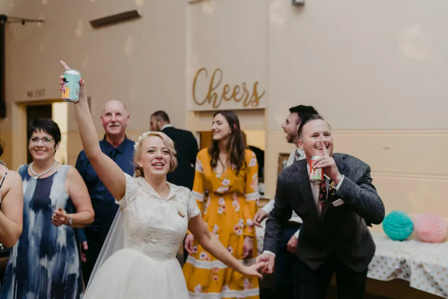 Bride raising a drink while holding the groom's hand among cheering guests.