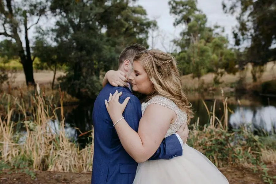 Bride hugging a man in a blue suit beside a garden pond.