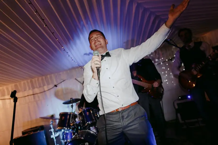 Man singing into a microphone with one arm raised beside the band.
