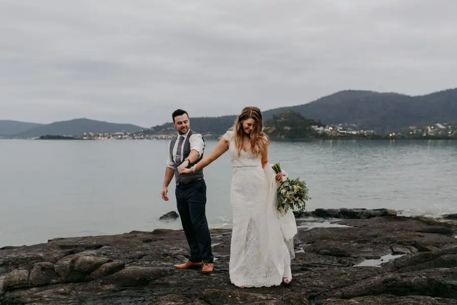 Couple holding hands on shoreline rocks with hills across the water.