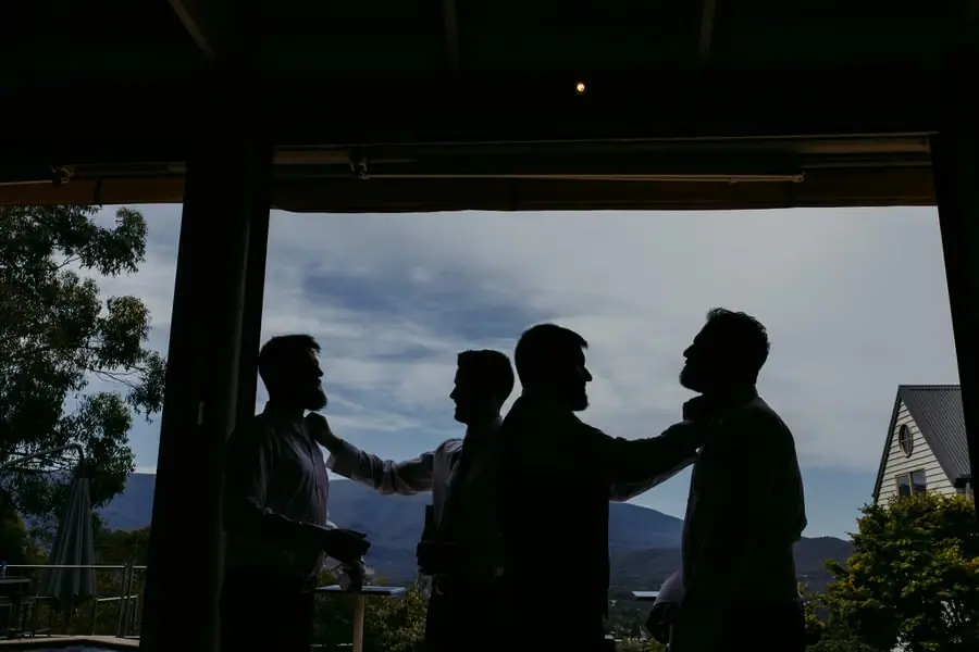 Silhouetted men adjusting each other's clothing beside wide windows.