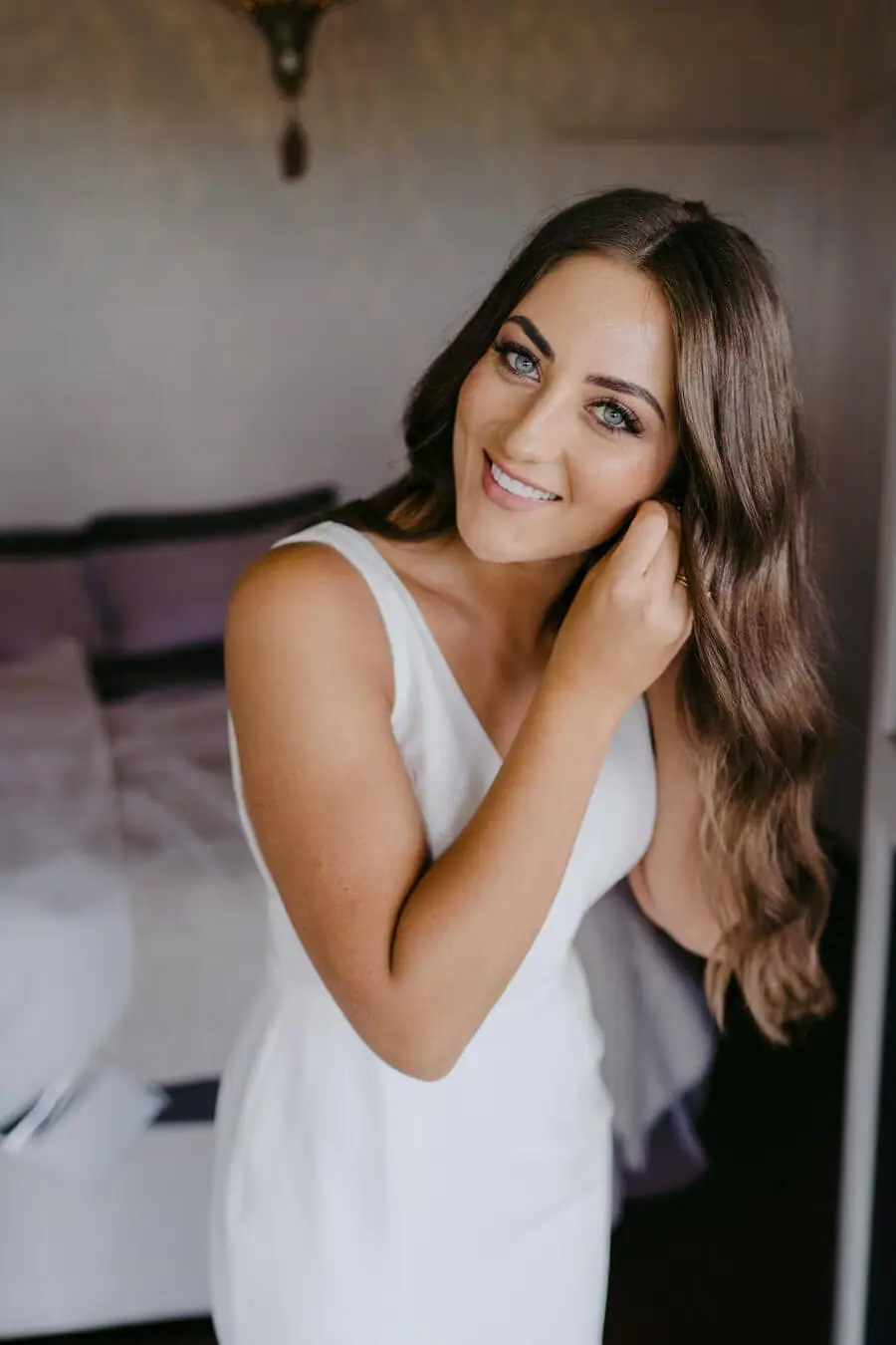 Bride smiling and adjusting an earring in a bright doorway.