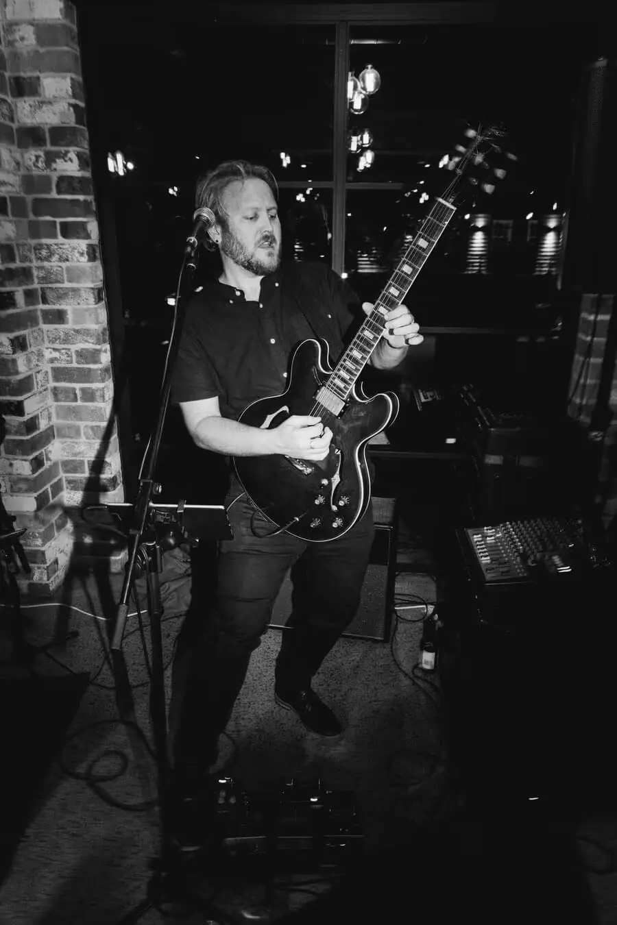 Black-and-white photograph of a guitarist performing beside a brick pillar.