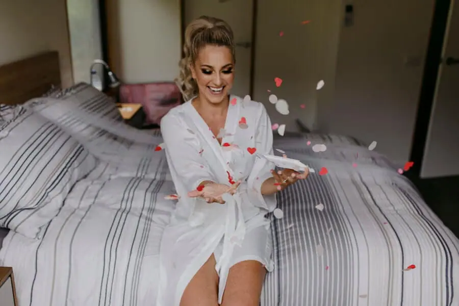 Bride in a robe tossing flower petals while sitting on a bed.