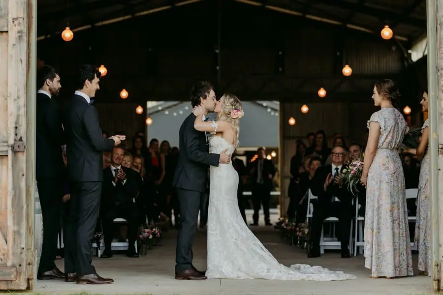 Bride and groom kissing at the front of a rustic indoor ceremony.