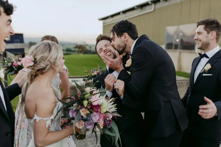 Men sharing an embrace and a cheek kiss beside the bride outdoors.