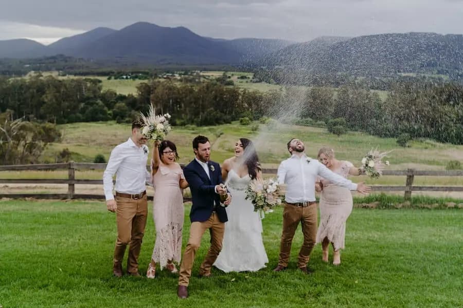 Wedding party laughing while a bottle sprays sparkling wine over the lawn.
