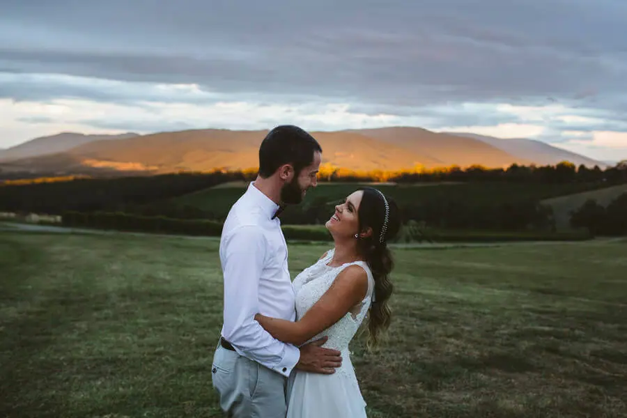 Couple embracing on a lawn beneath distant sunset-lit mountains.