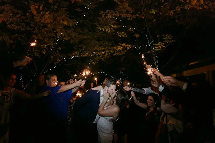 Couple kissing beneath sparklers held by guests at night.