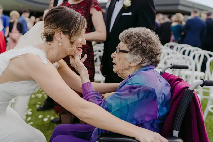 Bride leaning toward an older woman seated in a wheelchair.