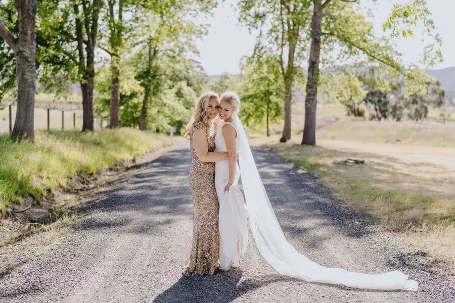 Two brides embracing on a tree-lined country road.