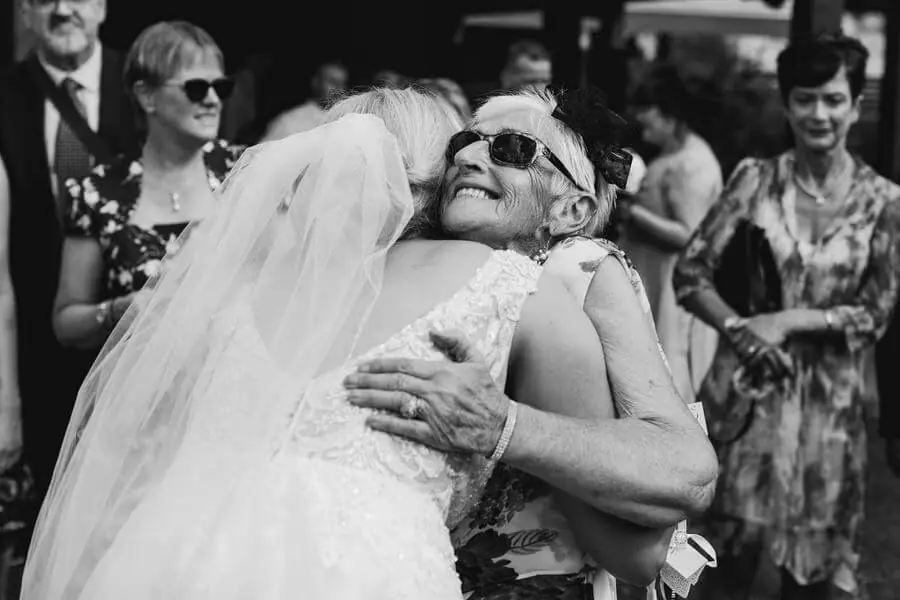 Black-and-white photograph of a woman in sunglasses hugging the bride.