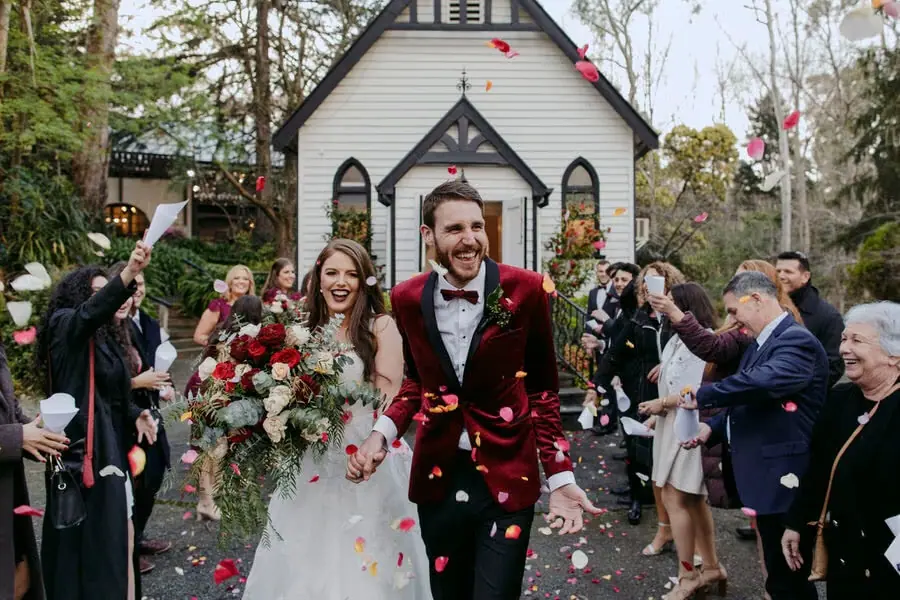 Couple walking through red petals outside a small chapel.
