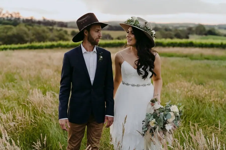 Couple wearing broad-brimmed hats and holding hands in tall grass.