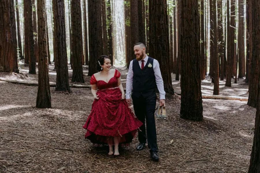 Woman in a red dress walking hand in hand with a man through tall forest trees.