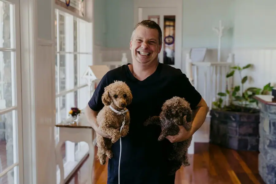 Man laughing while holding two small curly-haired dogs indoors.