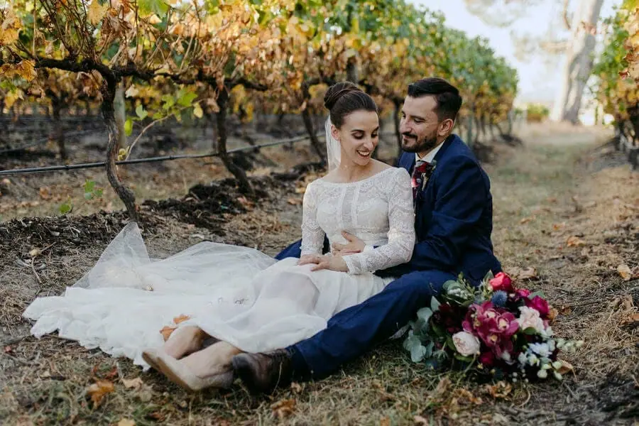 Bride and groom sitting among fallen leaves beside vineyard rows.