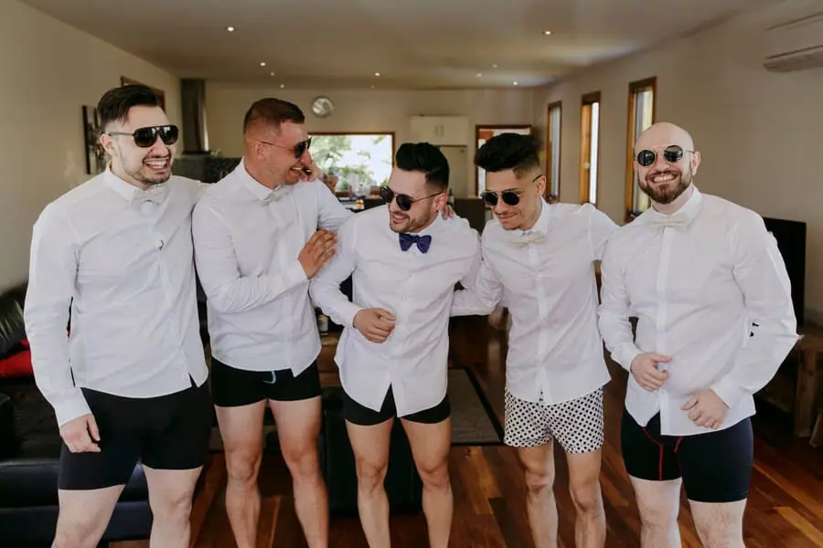 Five men in white shirts and underwear laughing during preparations.