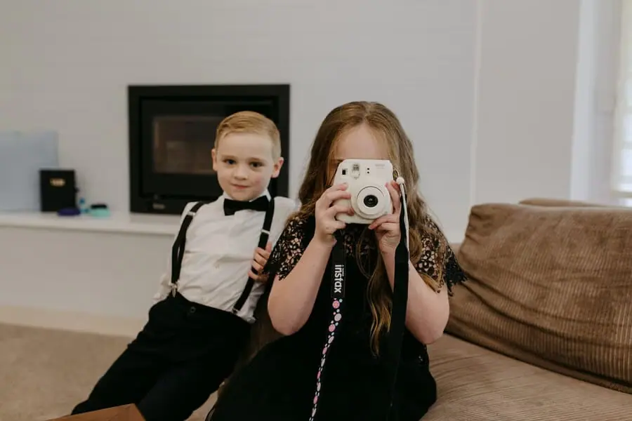Two children sitting on a sofa as a girl holds a small instant camera.
