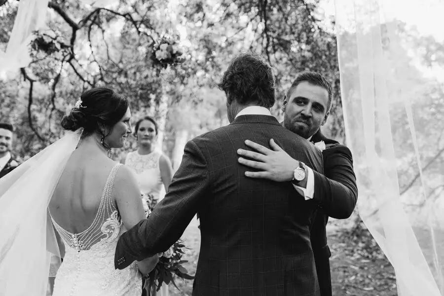 Black-and-white photograph of a groom hugging a man beside the bride at a woodland ceremony.