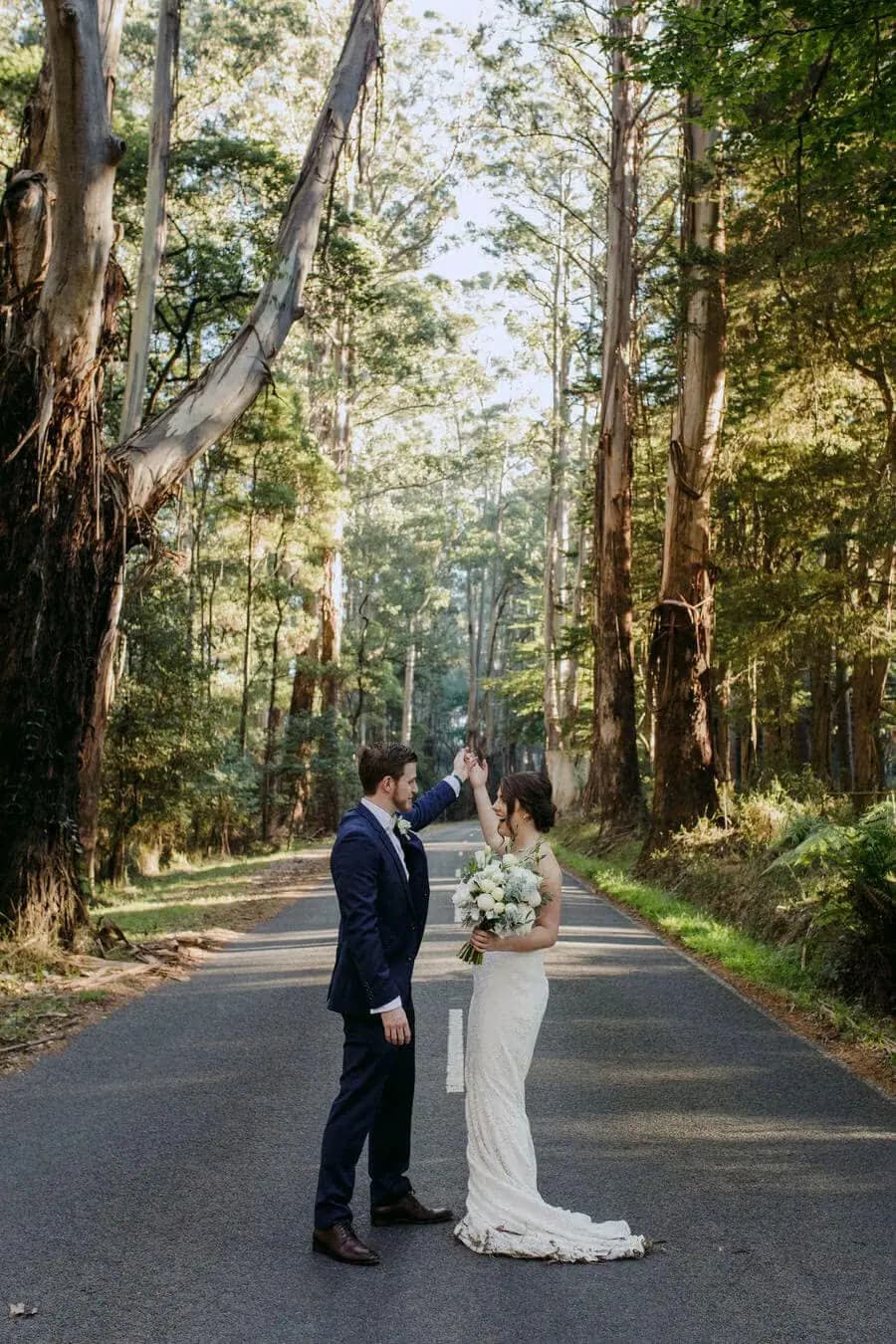 Bride and groom standing together on a quiet road through tall trees.