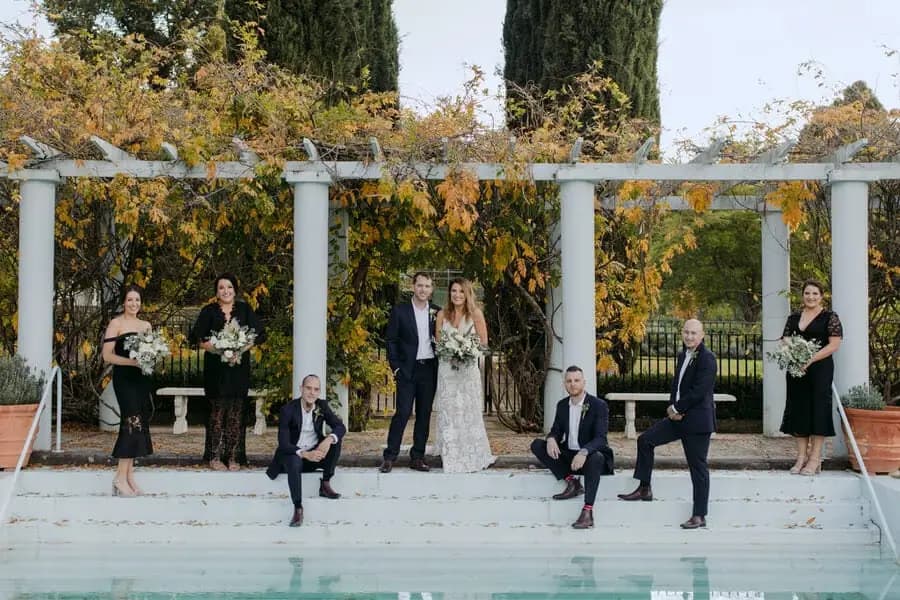 Wedding party arranged beside a pool beneath an autumn-covered pergola.