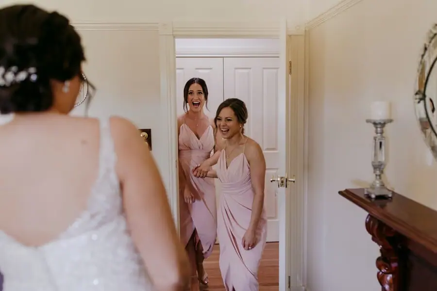 Women in pink dresses reacting to the bride inside a bedroom.