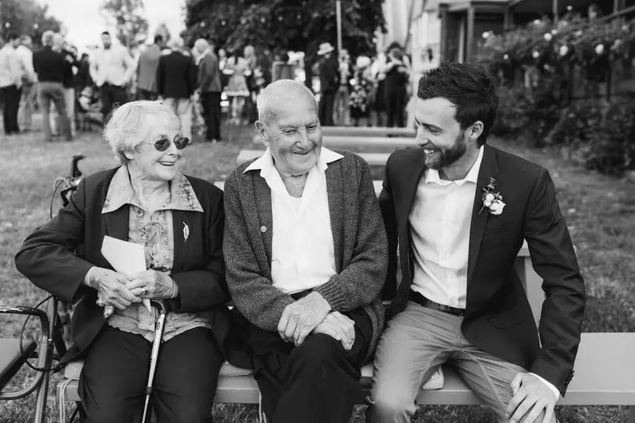 Black-and-white photograph of a groom sitting and smiling with two older guests.