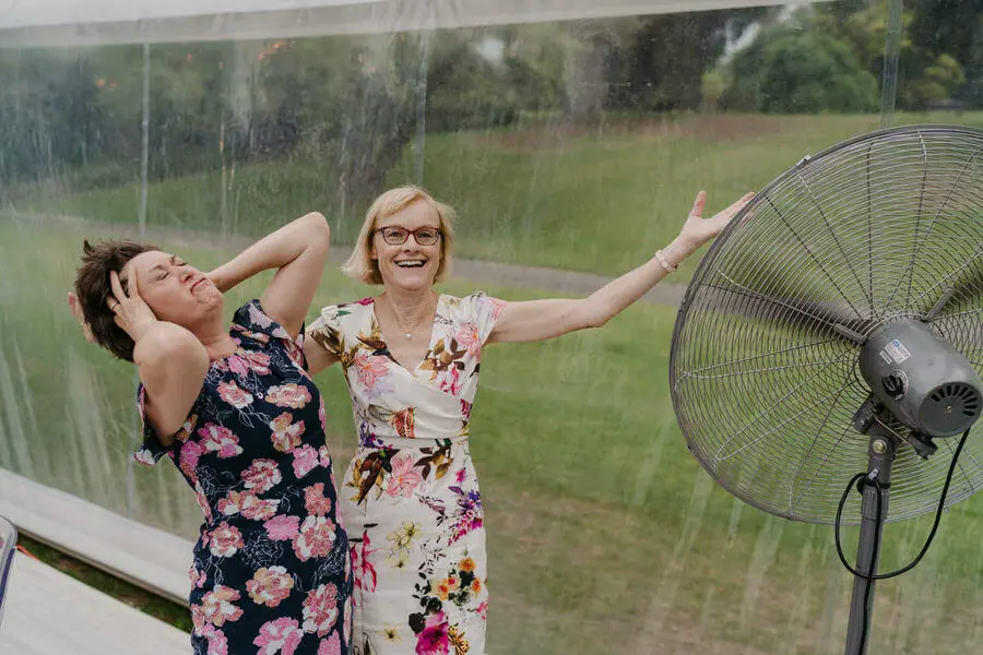 Two women laughing beside a large floor fan.