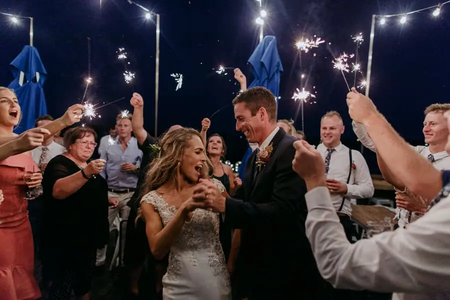 Bride and groom smiling among guests holding sparklers at night.