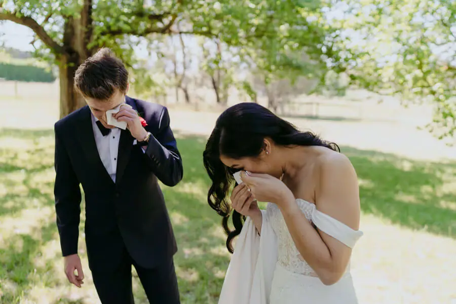Bride and groom each wiping their face with a tissue outdoors.