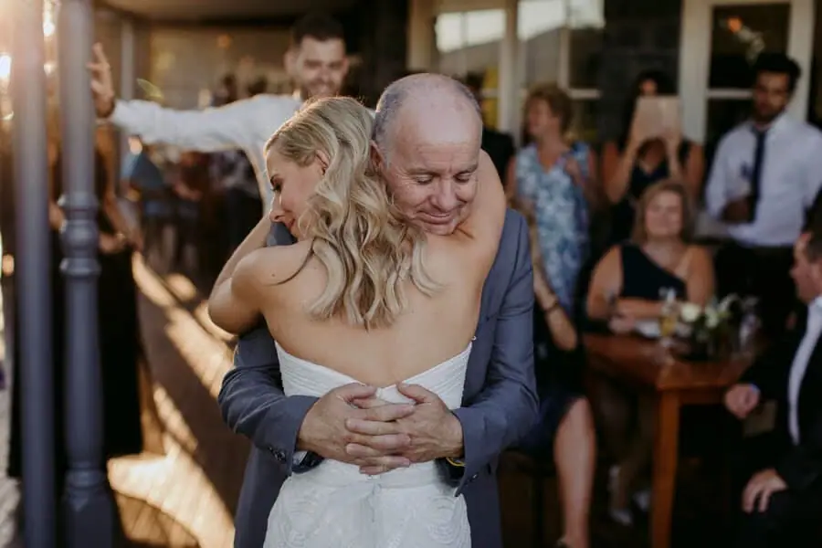 Bride hugging a man as guests watch inside the reception.