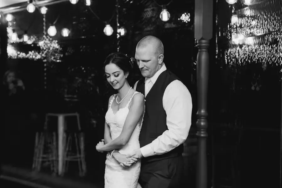 Black-and-white photograph of a man embracing a woman from behind beneath lights.