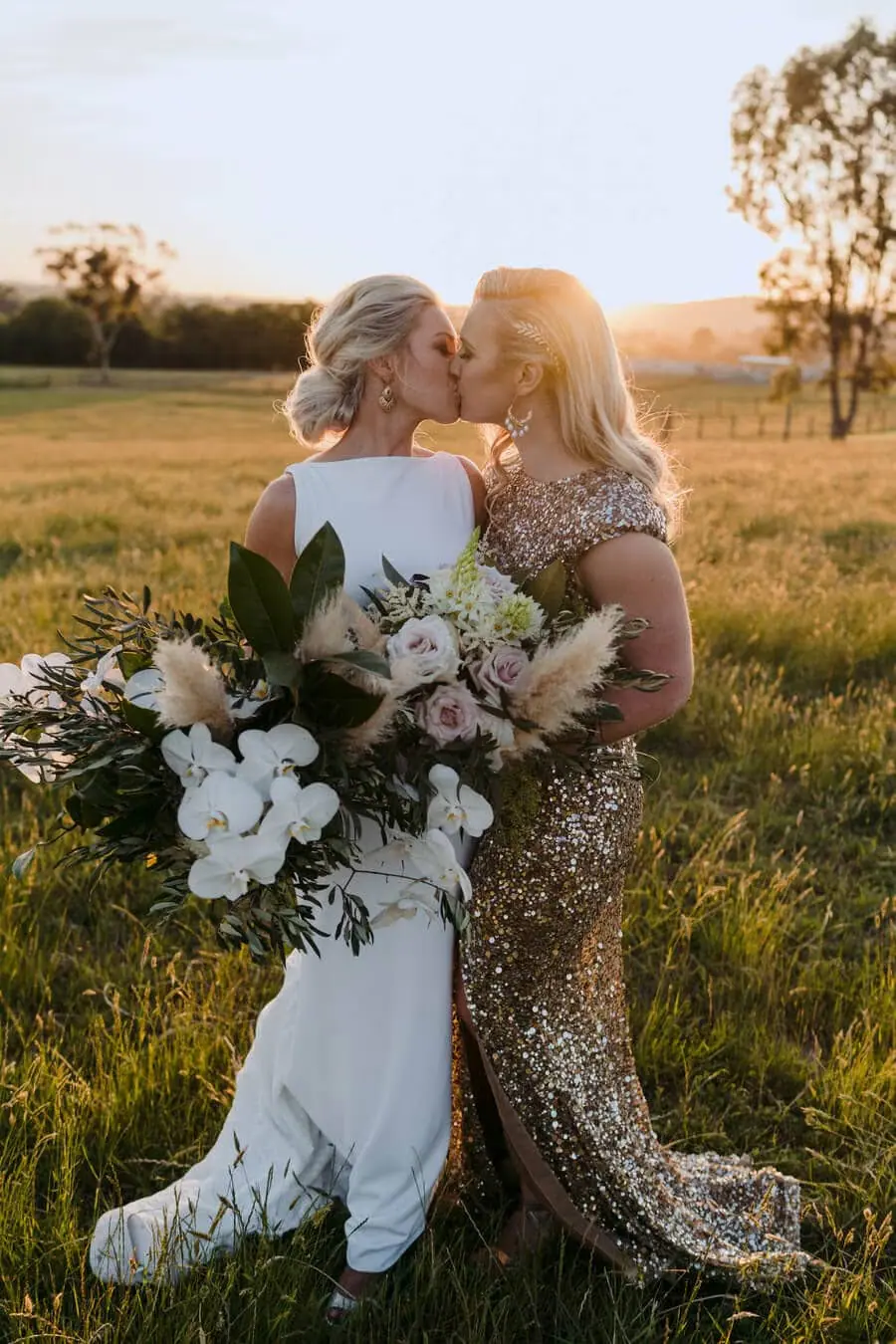Two brides kissing in a sunlit field with white flowers.