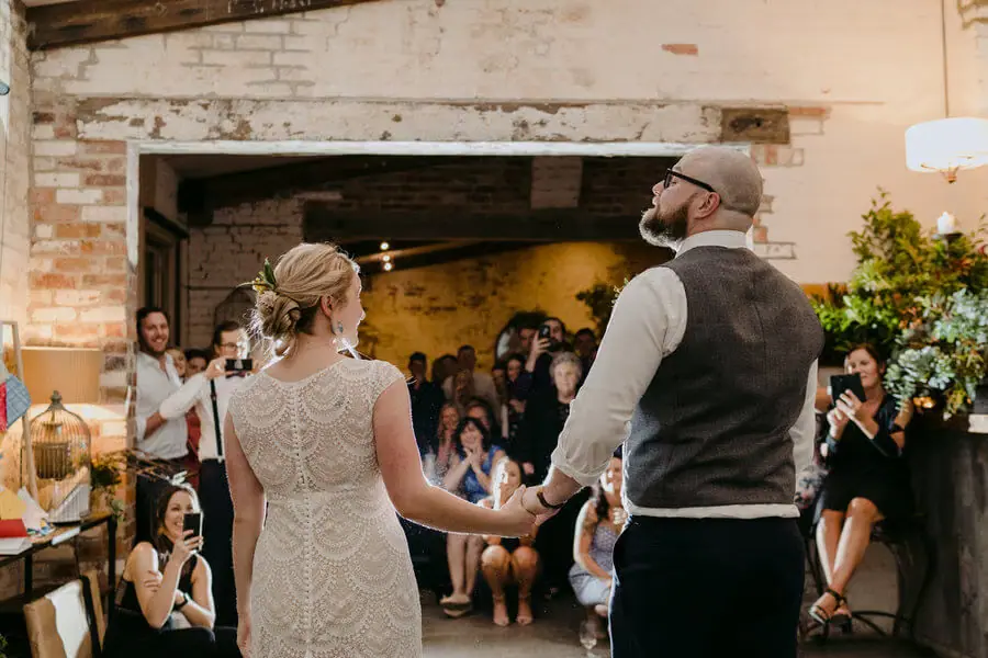 Bride and groom holding hands before watching guests inside a rustic reception room.