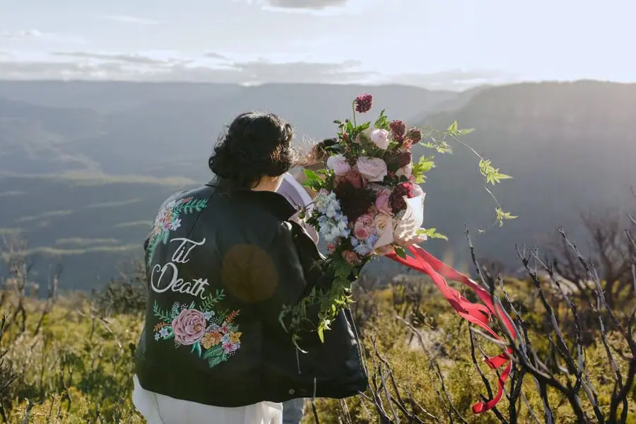 Bride holding a colourful bouquet and wearing a decorated jacket overlooking hills.